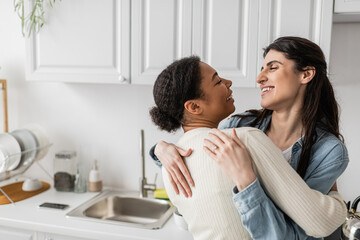 overjoyed lesbian woman smiling and hugging positive multiracial girlfriend with curly hair.