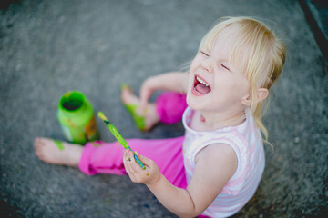 A cheerful cute girl draws with green paints on her leg, the foot of her body. Toddler girl plays on the street. Real sincere moments from the life of childhood. The young artist paints on the body. F