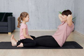 Fototapeta na wymiar family and sport concept - young mother and cute little daughter doing sport exercises for abdominal muscles together at home