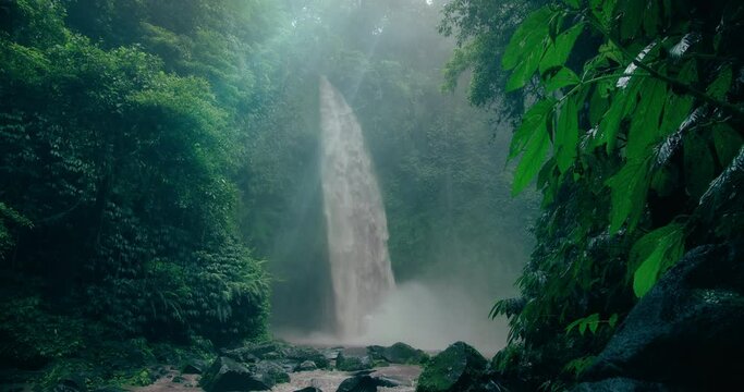 Nungnung waterfall lost between old green tropical trees and vegetation. Thick jungle after rain. Wet foliage shining in dim light of dense woods. Mystic rainforest.