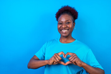 Young african american woman isolated on a blue background smiling and heart gesture, studio shoot