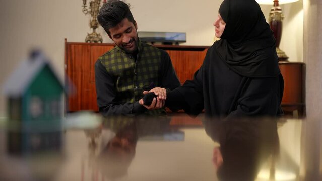 Satisfied happy Middle Eastern man talking with Caucasian Muslim woman in hijab at home. Portrait of loving young couple in living room indoors