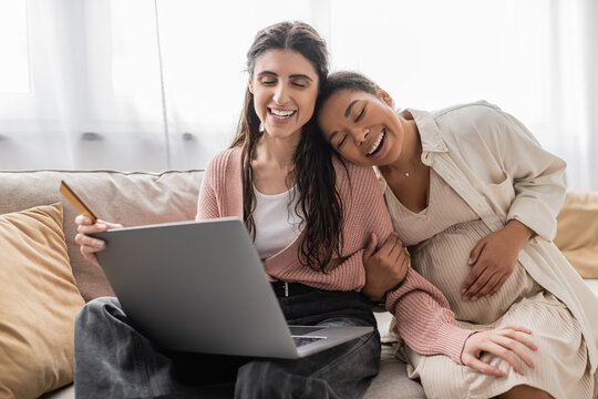 happy lesbian woman holding credit card while doing online shopping near pregnant multiracial partner.