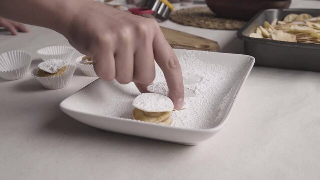 zoom out of hand removing leaf litter alfajores from the powdered sugar sifting area, then puts it in a white capsule on a table with pastry items. Home environment with natural light
