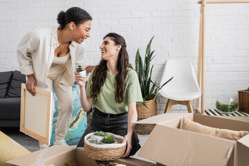 happy lesbian woman showing tiny green plant to multiracial girlfriend holding paintings in new...