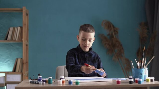 Caucasian guy schoolboy sits at home in casual clothes draws a picture with a brush on canvas. Children's education, entertainment, hobby,