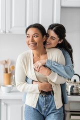 cheerful lesbian woman hugging positive multiracial girlfriend in modern kitchen.