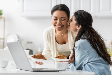 Fototapeta premium happy lesbian woman kissing cheek of cheerful multiracial girlfriend with croissants.