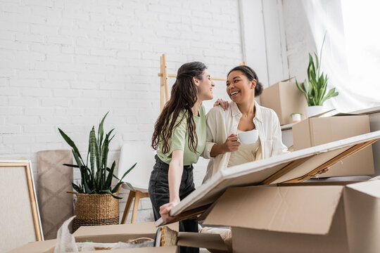 joyful interracial lesbian couple looking at each other while unpacking painting from box in new house.