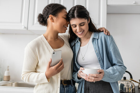 joyful multiracial woman holding smartphone and hugging lesbian partner with cup of coffee.