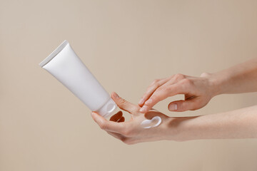 Fragile hands of a woman hold a white mockup tube of facial cream and apply moisturizer to her skin on a beige isolated background. Image for your design.