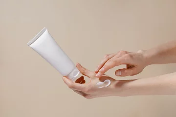 Poster Fragile hands of a woman hold a white mockup tube of facial cream and apply moisturizer to her skin on a beige isolated background. Image for your design. © Daryna 