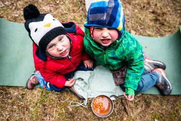 Two children smiling while eating a meal in front of a tent at a campsite.