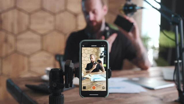 Young bearded charismatic Caucasian man is recording video review on new flagship smartphone model and using mobile phone mounted on tripod instead of camera sits at table in home office.