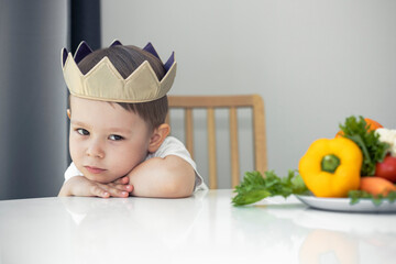 Sad little boy with a plate of fresh vegetables at the table. The child in the crown hates to eat...