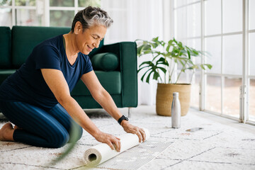 Getting ready for a pilates session, elderly woman rolls out her fitness mat at home