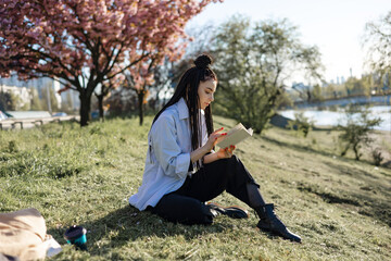 Young woman with dreadlocks reads book in spring park.