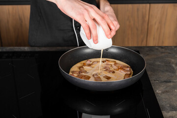A Cook in a black uniform in the kitchen with ingredients prepared for cooking. Chef making an omelet with bacon for breakfast.
