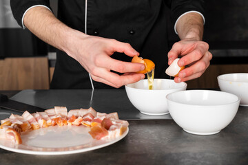A Cook in a black uniform in the kitchen with ingredients prepared for cooking. Chef making an omelet with bacon for breakfast, separating the yolk from the egg white.