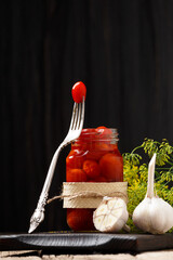 Pickled tomatoes in an open jar, one tomato on a fork, garlic close-up on a dark wooden background.