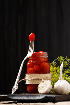Fototapeta Pickled tomatoes in an open jar, one tomato on a fork, garlic close-up on a dark wooden background.