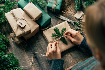 An unrecognizable woman is wrapping a Christmas gift in craft paper and a fir branch.