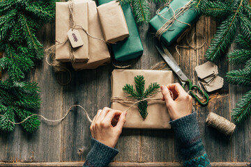 An unrecognizable woman is wrapping a Christmas gift in craft paper and a fir branch.
