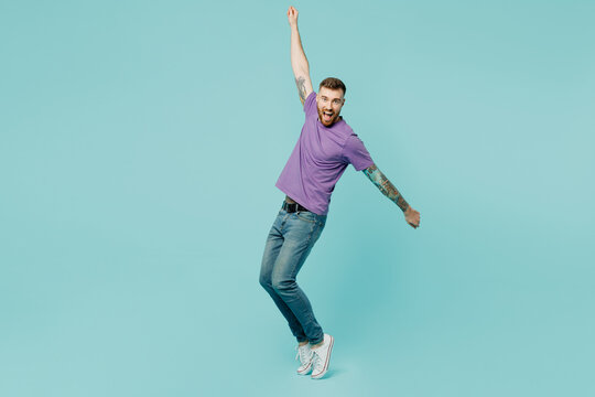 Full body european young man he wears purple t-shirt stand on toes with outstretched hands leaning back dancing isolated on plain pastel light blue cyan background studio portrait. Lifestyle concept.