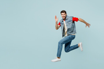 Full body side view fun cool cheerful young man he wears denim vest red t-shirt casual clothes jump high run fast isolated on plain pastel light blue cyan background studio portrait Lifestyle concept