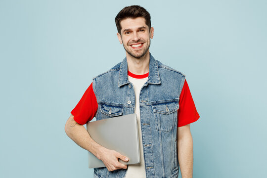 Young fun happy student IT man he wear denim vest red t-shirt casual clothes holding closed laptop pc computer isolated on plain pastel light blue cyan background studio portrait. Lifestyle concept
