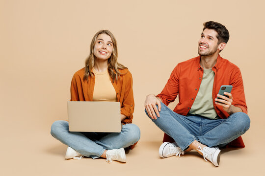Full body young couple two friends family IT man woman wearing casual clothes together sit hold work on laptop pc computer use mobile cell phone isolated on pastel plain light beige color background.