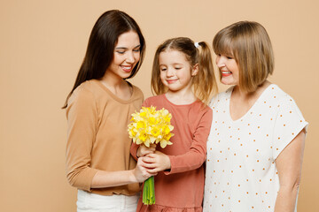 Fototapeta na wymiar Happy lovely women wear casual clothes with child kid girl 6-7 years old. Granny mother daughter gift giving bouquet of daffodil flowers isolated on plain beige background. Family parent day concept.