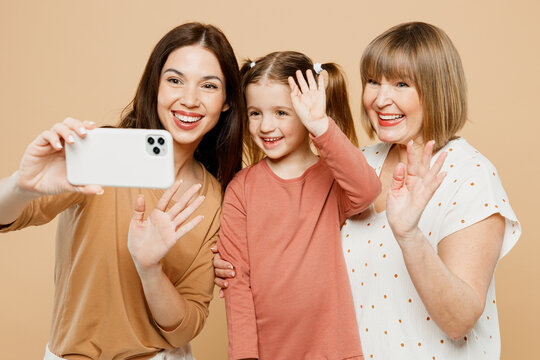 Happy positive women wear casual clothes with child kid girl 6-7 years old. Granny mother daughter doing selfie shot on mobile cell phone isolated on plain beige background. Family parent day concept.