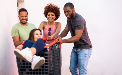 A group of 3 multiracial millennial youths have a good time pushing a girl inside a shopping cart outside the street. Concept of fun among young people, carefree, end of exams, university parties. - obrazy, fototapety, plakaty