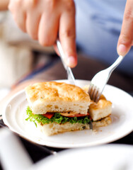 Person holding a sandwich. Person splits sandwich by fork and knife. Bread with meat and cabbage on white plate. 