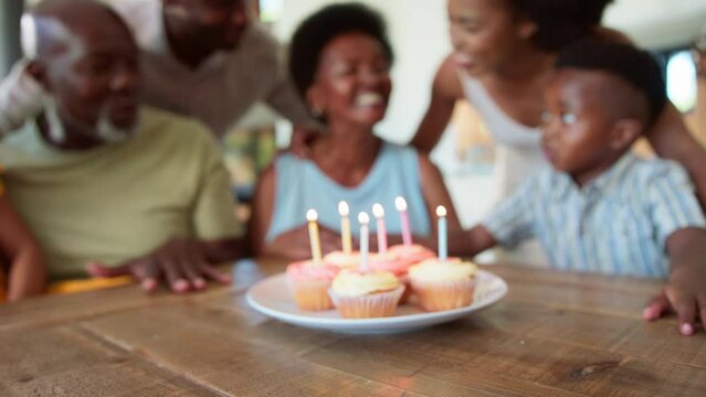 Multi-generation family sitting around table at home celebrating grandmother's birthday singing happy birthday before she blows out candles on birthday cake or cupcakes together- in slow motion