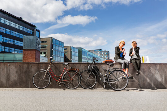 Two women with bicycles sitting on wall and talking