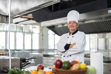 Confident senior Asian chef man in dress uniform standing and arms crossed with various vegetables in kitchen restaurant. Cooking and food concept