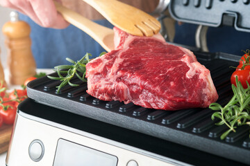 Raw prime steaks on electric grill. Chef male hand holding steak Ribeye on modern electric grill with rosemary, cherry tomato and pepper. Cooking process in modern kitchen. Top view, copy space.