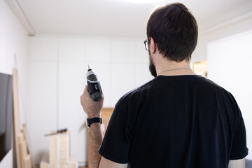 Back view a man with a smartwatch with glasses holding a drill or electric screwdriver the background of a room before assembling furniture. Tools, accessories, installing furniture, repairing a house