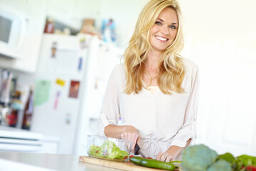 Preparing a salad for dinner. Attractive young blonde woman smiling while chopping fresh vegetables at home.