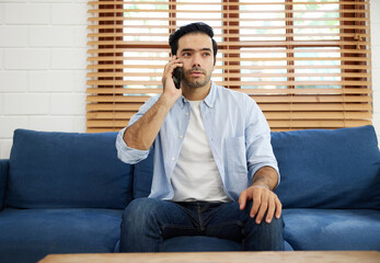 Fototapeta premium young handsome man sitting on sofa and talking by smartphone at home
