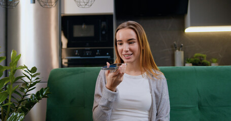 Beautiful teenage girl sitting on sofa at home holding cellphone talking on speaker phone, leaves audio message. Sound recognition in mobile assistance app sets reminder on phone at home use digital