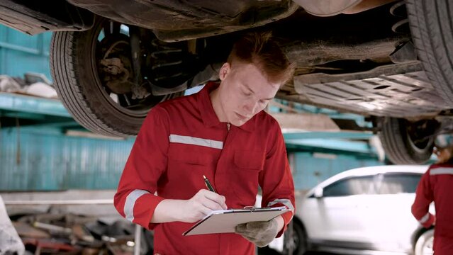 Close-up car mechanic, wear work clothes for work safety prevent fouling, man mechanic holding car condition checkbook, wearing gloves rocking car suspension check at point that needs be repaired.
