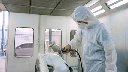 worker wearing PPE suit over head wear goggles mouth mask, standing in paint room, repainting car that is not airy, mechanic uses spray machine slowly spray paint onto car's surface.