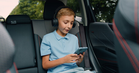 Good-looking relaxed blond teen boy singing songs to music from his phone playlist using earphones while driving in a car