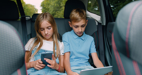 Cute relaxed teen boy and girl sitting on car's backseat and use tablet pc and smartphone during trip,front view