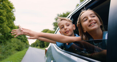 Attractive satisfied smiling teen brother and sister put their heads out of auto window,waving...