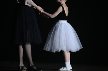 A young ballerina with a mentor studying in lesson standing in a black classroom holding hands