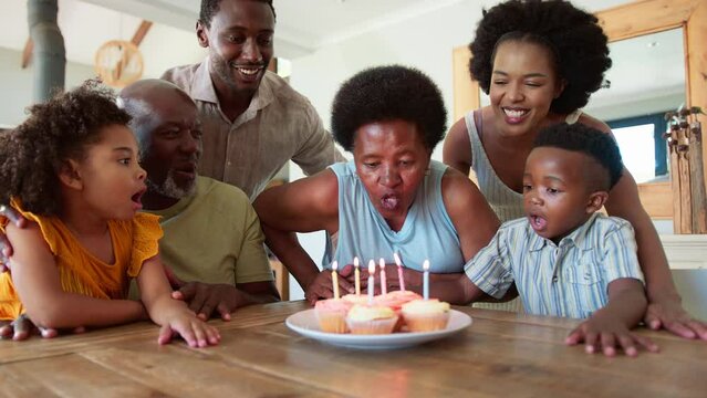 Multi-generation family sitting around table at home celebrating grandmother's birthday singing happy birthday before she blows out candles on birthday cake or cupcakes together- in slow motion